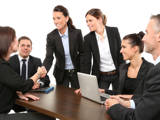 Six people in a meeting room with two people standing. One standing person is shaking hands with a seated person.